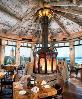 A rustic restaurant interior with a high, wooden ceiling and large panoramic windows offering stunning views of a lake and forest. The space features a central, enclosed fireplace, surrounded by dining tables set with white linens, brown chairs, and wooden floors. - Hausion