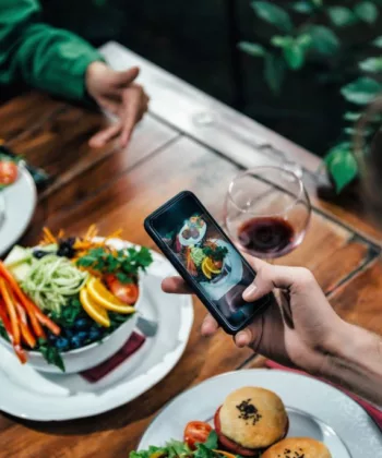 A person photographing a plate of food with a smartphone at a wooden table. The table is set with several colorful dishes, including salads and burgers, and a glass of red wine. Another person's hands are visible in the background, gesturing as they talk. - Hausion