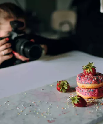 A person is photographing a stack of three colorful, sprinkled donuts topped with strawberries on a marble surface. Several strawberries and sprinkles are scattered around the donuts. In the background, there are blurred objects, adding depth to the image. - Hausion