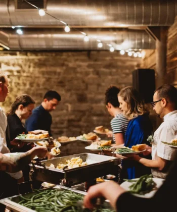 A group of people are standing in line at a buffet in a rustic setting with exposed brick walls and wooden panels. They are serving themselves food from chafing dishes filled with various dishes. String lights hang from the ceiling, creating a warm ambiance. - Hausion