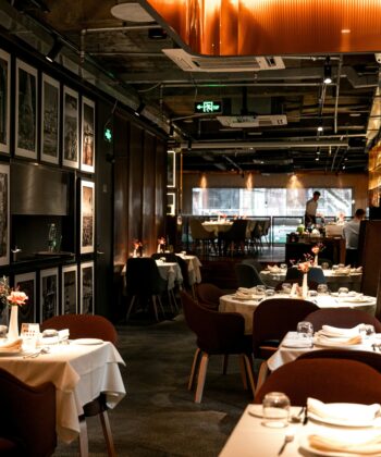 A modern, dimly-lit restaurant with elegant decor and neatly set tables. Several framed black and white pictures adorn the left wall. The dining tables are covered with white tablecloths, and soft lighting creates a warm ambiance. A person is visible in the background. - Hausion