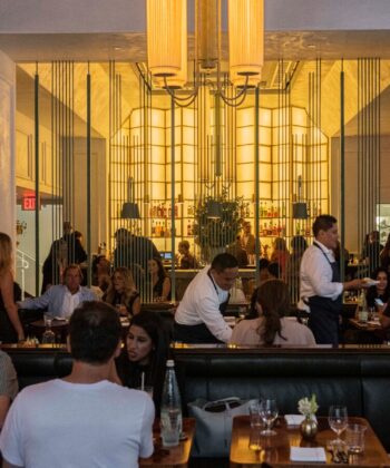 A lively restaurant scene with people dining and socializing. The modern interior features a warm yellow-lit bar at the back, large vertical light fixtures, and a mix of seated and standing patrons. Servers bustle around attending to customers.

. - Hausion