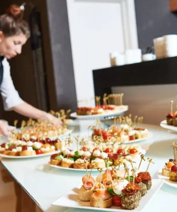 A waitress in a black apron and white shirt arranges several plates of assorted appetizers and hors d'oeuvres on a transparent table. The appetizers include skewers with various ingredients like tomatoes, cheese, and meats, presented on small, decorated picks. - Hausion