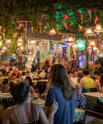 A lively outdoor restaurant is bustling with people dining under twinkling string lights and decorative hanging lamps. Colorful buntings zigzag overhead, and greenery drapes from the ceiling. Patrons sit at closely packed tables, engaged in conversation and enjoying the atmosphere. - Hausion