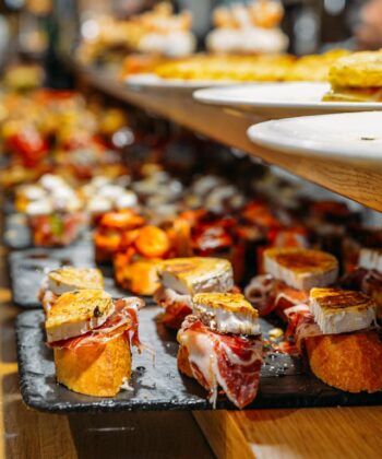 A selection of gourmet tapas on a wooden counter, including small toasted slices of bread topped with various ingredients such as cheese, cured meats, and vegetables. The tapas are arranged on black stone plates with a blurred background. - Hausion