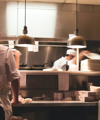 A busy restaurant kitchen with chefs in white uniforms working. In the foreground, a chef is seen from the back organizing ingredients. In the background, other chefs are blurred in motion, plating dishes under industrial lights. Shelves holding plates are visible. - Hausion