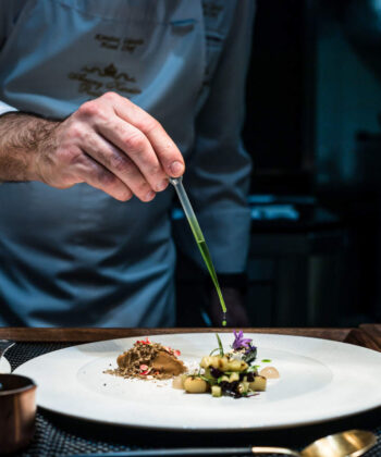 A chef in a white coat carefully garnishes a plated dish with precision using tweezers. The dish is artistically presented with various colorful ingredients on a white plate. A small saucepan and utensils are visible on the table. The setting appears to be a professional kitchen. - Hausion