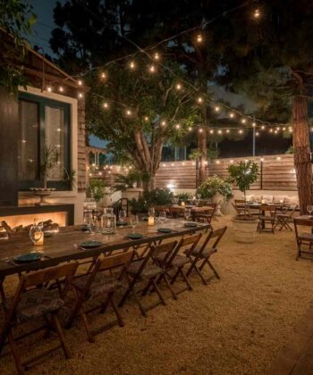 An outdoor dining area is set at night, featuring wooden tables and chairs on a gravel surface. String lights are hung across trees and the surrounding fence, creating a warm, inviting ambiance. The tables are set with plates, glasses, and candles, ready for guests. - Hausion