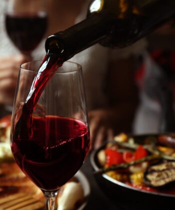 A close-up of red wine being poured into a glass, set against a table filled with various dishes, including grilled vegetables and cheese. The background is slightly blurred, showing a person seated and holding a glass. The ambiance appears warm and cozy. - Hausion