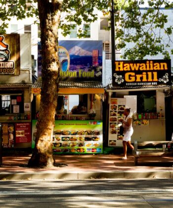 A street scene featuring several food stalls lined up side by side, including "Don Pedro Mexican Food," "Hawaiian Grill," and "Veli Thai Food." Two people sit on a bench in front of the stalls, while two others stand nearby. Trees provide shade over the street and vendors. - Hausion