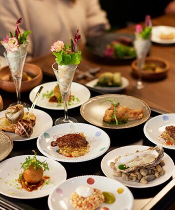 A table is filled with an assortment of elegantly presented dishes, including seafood, meat, and vegetarian options. The plates are meticulously arranged, some with vibrant garnish. Several people are seated around the table, enjoying the meal in a warm, inviting atmosphere. - Hausion