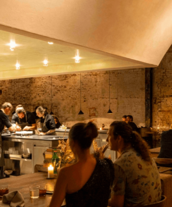 A cozy, dimly-lit restaurant with exposed brick walls. Diners sit at wooden tables, engaged in conversation, while chefs in black attire prepare meals behind a sleek, well-lit counter. Small hanging lights create a warm and intimate ambiance. - Hausion