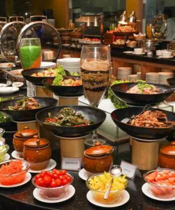 A lavish buffet spread featuring a variety of dishes in large black bowls and jars. The selection includes fresh salads, assorted vegetables, condiments, sauces, meats, and grains. In the background, chafing dishes and plates are arranged for hot items. - Hausion