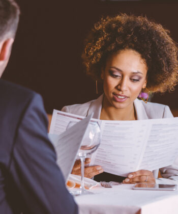 A woman with curly hair and a man in a suit are sitting at a table in a dimly lit restaurant, looking at menus. The table is set with water glasses, a folded napkin, and a smartphone. Red chairs and a dark background are visible. - Hausion