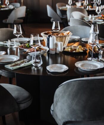 A round dining table set for a meal in an elegant restaurant. The table features several white plates, glasses, napkins, and silverware arranged neatly. A basket of bread and various dishes are placed in the center. Grey cushioned chairs surround the table. - Hausion