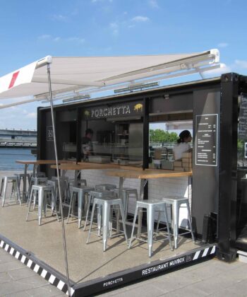 A modern outdoor food stall named "Porchetta" set up in a shipping container, offering shaded bar seating with metal stools. Two staff members are inside, serving food. The stall is located by a waterfront with a view of a bridge in the background. - Hausion