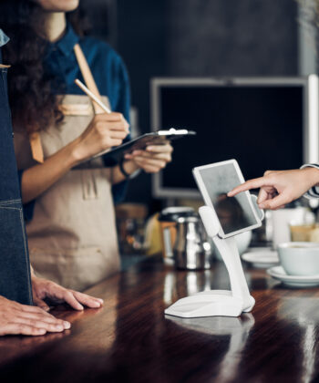 Two cafe staff members, one holding a clipboard, stand behind a wooden counter. A customer is using a touchscreen device on the counter. Drinkware, such as cups and saucers, are visible in the background. - Hausion