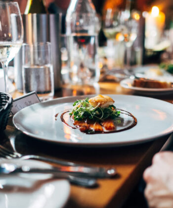 A close-up of a gourmet meal served on a white plate, featuring a small portion of food garnished with greens and sauce. The table is elegantly set with wine glasses, candles, and silverware, suggesting a fine dining experience. - Hausion