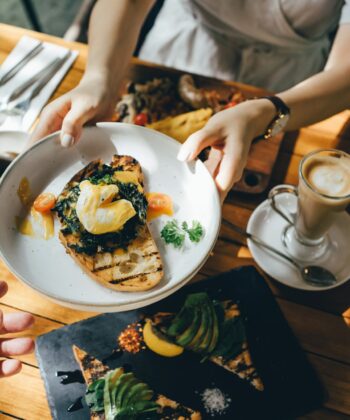 A person hands a plate of toast topped with poached eggs, greens, and tomatoes to another person's outstretched hand over a wooden table. The table is set with various dishes, including a latte, a glass of orange juice, and avocado toast. - Hausion