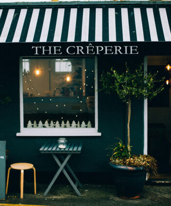 A cozy crepe cafe with a black and white striped awning labeled "The Crêperie." Outside, there's a chalkboard menu on a small wooden stand, a table with two chairs, potted plants, and festive lights around the window. A larger menu is on the right wall. - Hausion