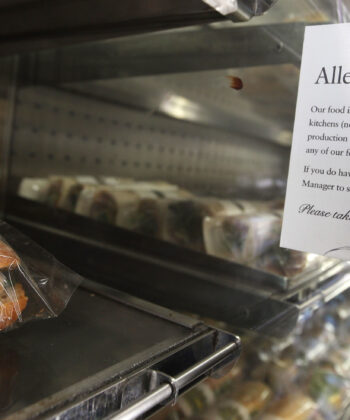 A display case with sandwiches is shown. Next to the sandwiches, there is a sign that reads, "Allergies? Our food is freshly prepared in small, busy kitchens (not in a factory with segregated production lines) so we really can't guarantee any of our food is allergen-free. - Hausion