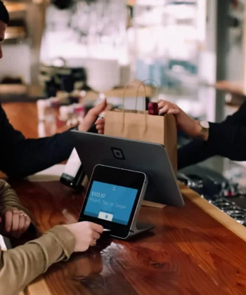 A barista hands a paper bag to a customer across the counter in a café. The customer is making a payment on a digital point-of-sale terminal, while another customer stands beside them. The scene shows a friendly transaction in a well-lit establishment. - Hausion