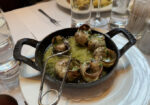 A dish of escargot served in a black cast-iron pan, featuring several cooked snails in shells swimming in a garlic and herb butter sauce. A special snail tong is placed on the edge of the pan. The table includes water glasses, cutlery, and other dishes in the background. - Hausion