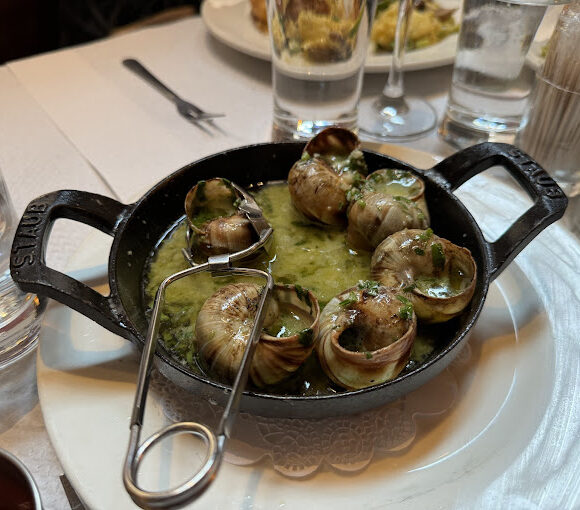 A dish of escargot served in a black cast-iron pan, featuring several cooked snails in shells swimming in a garlic and herb butter sauce. A special snail tong is placed on the edge of the pan. The table includes water glasses, cutlery, and other dishes in the background. - Hausion