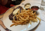 A plate of food featuring a grilled steak topped with herb butter, a side of golden French fries, a small metal dish of mayonnaise, and a small metal dish of ketchup. In the background, a person in a red shirt is holding a wine glass. - Hausion