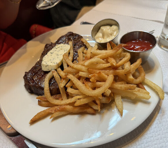 A plate of food featuring a grilled steak topped with herb butter, a side of golden French fries, a small metal dish of mayonnaise, and a small metal dish of ketchup. In the background, a person in a red shirt is holding a wine glass. - Hausion