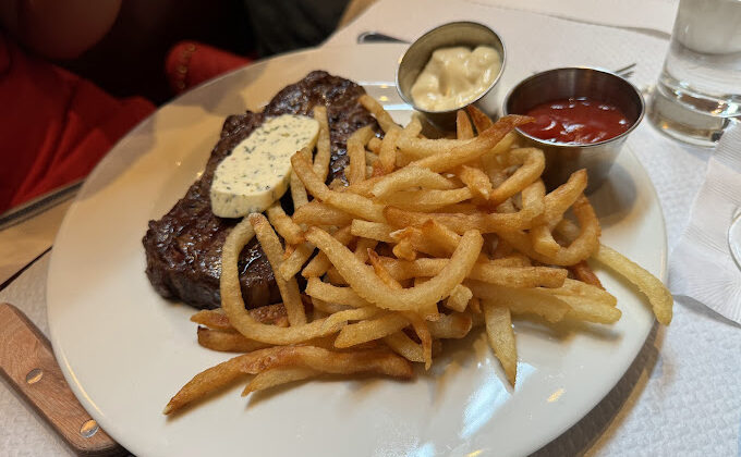 A plate of food featuring a grilled steak topped with herb butter, a side of golden French fries, a small metal dish of mayonnaise, and a small metal dish of ketchup. In the background, a person in a red shirt is holding a wine glass. - Hausion