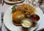 A plate featuring a cheeseburger topped with melted cheese, lettuce, and onions, accompanied by a generous serving of French fries. Two small metal dishes on the plate contain mayonnaise and ketchup. Forks and knives are placed around the plate. - Hausion