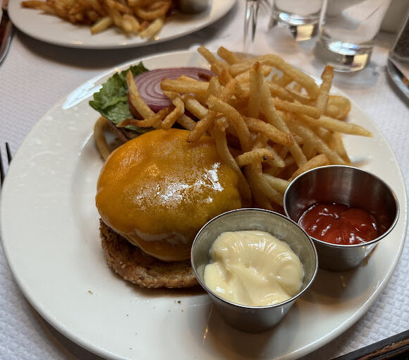 A plate featuring a cheeseburger topped with melted cheese, lettuce, and onions, accompanied by a generous serving of French fries. Two small metal dishes on the plate contain mayonnaise and ketchup. Forks and knives are placed around the plate. - Hausion