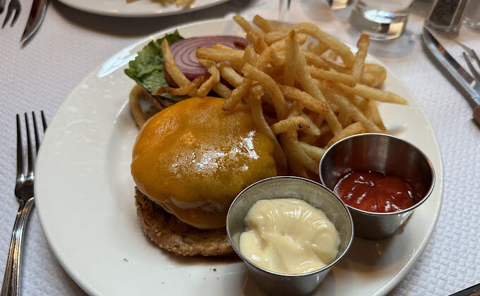 A plate featuring a cheeseburger topped with melted cheese, lettuce, and onions, accompanied by a generous serving of French fries. Two small metal dishes on the plate contain mayonnaise and ketchup. Forks and knives are placed around the plate. - Hausion