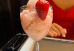 A glass of pink frozen beverage with a fresh strawberry garnish, placed on a table. Behind the glass, part of a person wearing a red shirt is visible. The blurred background suggests an indoor setting. - Hausion