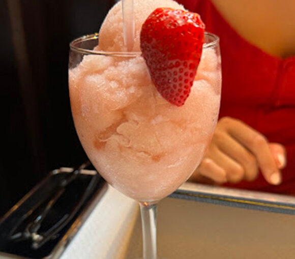 A glass of pink frozen beverage with a fresh strawberry garnish, placed on a table. Behind the glass, part of a person wearing a red shirt is visible. The blurred background suggests an indoor setting. - Hausion