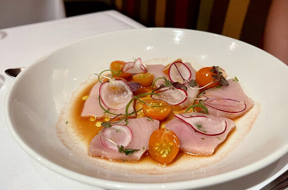 A white plate holds thin slices of raw fish, topped with halved cherry tomatoes, thinly sliced radishes, green herbs, and surrounded by a light brown sauce. The plate rests on a white tablecloth. - Hausion