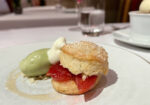 A dessert plate featuring a biscuit topped with a slice of fruit and a dollop of cream, accompanied by a scoop of green ice cream. In the background, there is a glass of water and another bowl with a scoop of vanilla ice cream on a white tablecloth. - Hausion