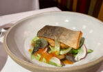 A carefully plated dish featuring a fillet of seared fish with crispy skin, resting on a bed of mixed greens, radishes, and herbs, garnished with orange fish roe. A glass of water is visible to the left side of the white tablecloth. - Hausion