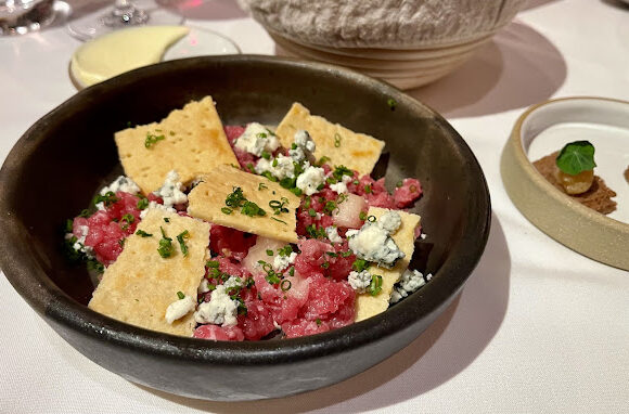 A gourmet dish is presented in a dark bowl, containing finely chopped, seasoned raw meat pieces, crumbled blue cheese, and scattered fresh herbs, topped with thin, crispy bread slices. A dish with small appetizers and a dipping sauce is in the background. - Hausion