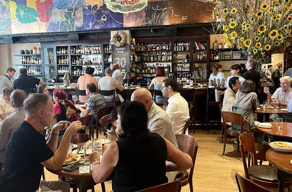 A bustling restaurant interior with patrons seated at wooden tables enjoying their meals. There is a bar in the background with a wide selection of bottles. The walls feature colorful artwork and a large vase with sunflowers decorates the right side of the image. - Hausion