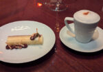 A white plate holds a rectangular dessert garnished with chocolate and nuts, next to a white cup of frothy cappuccino on a saucer, placed on a glossy wooden table. - Hausion