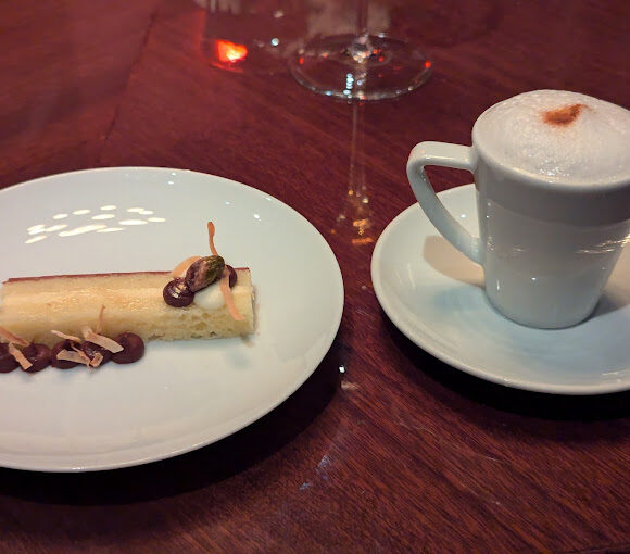 A white plate holds a rectangular dessert garnished with chocolate and nuts, next to a white cup of frothy cappuccino on a saucer, placed on a glossy wooden table. - Hausion