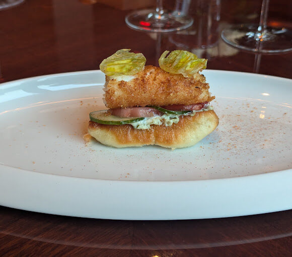 A gourmet appetizer featuring a small sandwich with a fried piece of fish, slices of cucumber, and a creamy spread. The sandwich is topped with two crispy herbs and is placed on a white oval plate. Stemmed glassware is in the blurred background. - Hausion