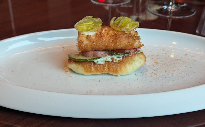 A gourmet appetizer featuring a small sandwich with a fried piece of fish, slices of cucumber, and a creamy spread. The sandwich is topped with two crispy herbs and is placed on a white oval plate. Stemmed glassware is in the blurred background. - Hausion