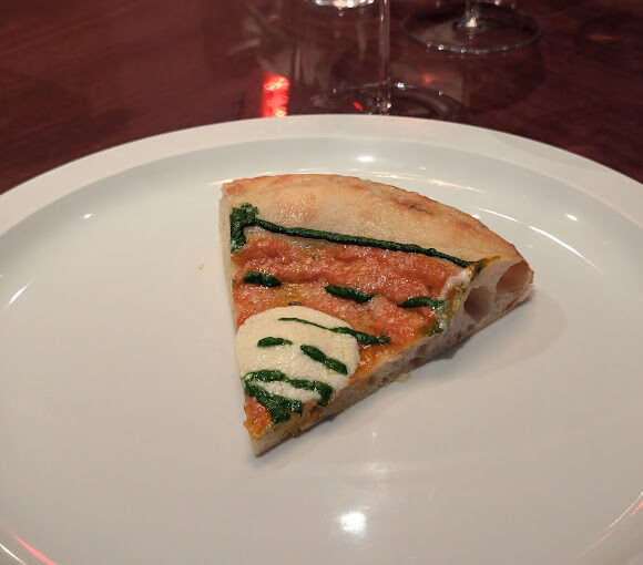 A single slice of pizza on a white plate. The pizza has a thin crust and is topped with a red sauce, a small amount of cheese, and drizzled with green sauce, possibly pesto. The plate is set on a wooden table with some glassware in the background. - Hausion