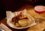 A plate of assorted fried seafood including shrimp, accompanied by a small bowl of dipping sauce and a wedge of lemon, sits on a wooden table. Beside the plate is a pink cocktail in a coupe glass. Napkins, cutlery, and another plate are also visible. - Hausion