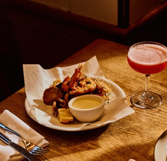 A plate of assorted fried seafood including shrimp, accompanied by a small bowl of dipping sauce and a wedge of lemon, sits on a wooden table. Beside the plate is a pink cocktail in a coupe glass. Napkins, cutlery, and another plate are also visible. - Hausion