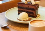 A slice of chocolate layer cake with a smooth chocolate glaze, topped with thin white sticks, is served on a green plate. The plate also holds a scoop of vanilla ice cream garnished with crumbs. Next to the plate is a cappuccino in a white cup with a spoon beside it. - Hausion