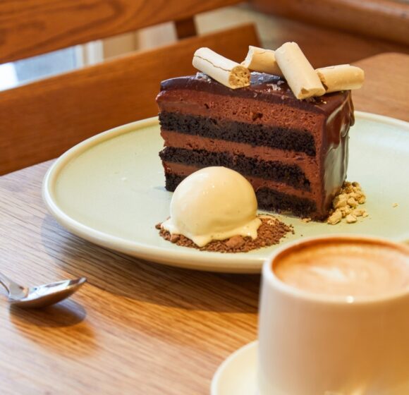 A slice of chocolate layer cake with a smooth chocolate glaze, topped with thin white sticks, is served on a green plate. The plate also holds a scoop of vanilla ice cream garnished with crumbs. Next to the plate is a cappuccino in a white cup with a spoon beside it. - Hausion